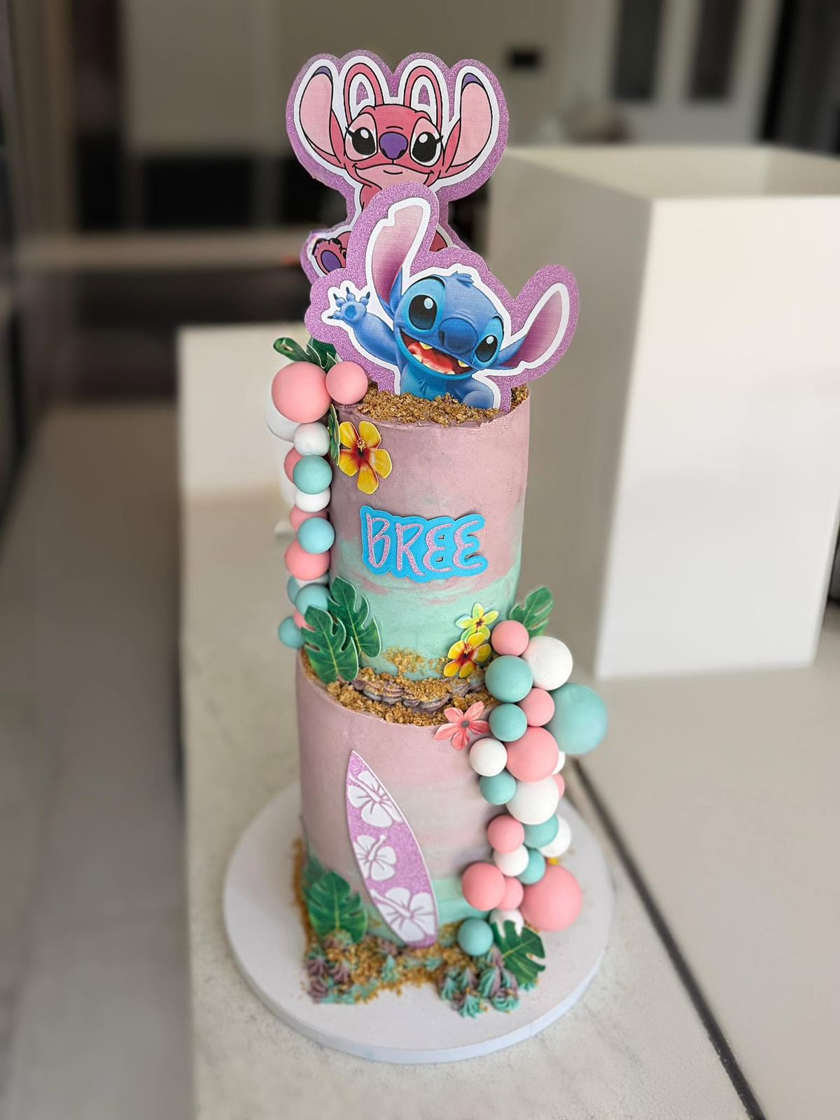 Kids’ character cake cake by Just In Cakes Co. Two tiers in pastel buttercream with pearl clusters, tropical leaves and a name.