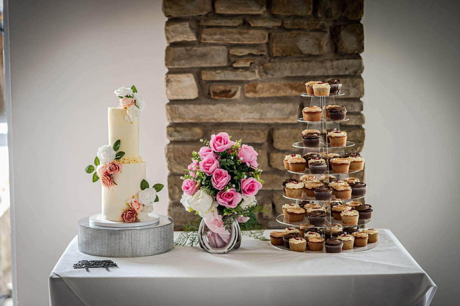 Wedding cake and cupcake tower cake by Just In Cakes Co. Two tiers with fresh roses and a cupcake tower, set up at the venue.