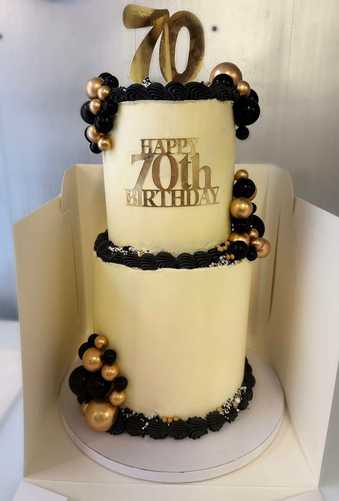 Black and gold 70th cake by Just In Cakes Co. Two tiers in ivory buttercream with black and gold pearls.
